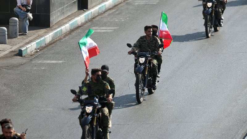 Men in uniform on motorcycles carrying large Iranian flags along a paved road.